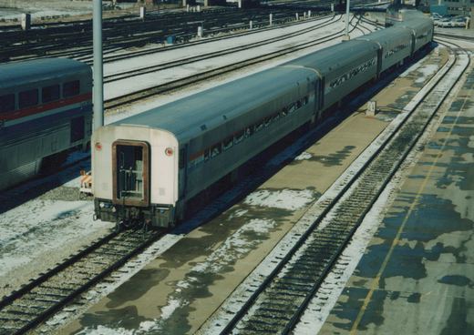 A high-angle view of an Amtrak passenger train at a snow-dusted station, showing the rear of a silver coach with a red and blue stripe.