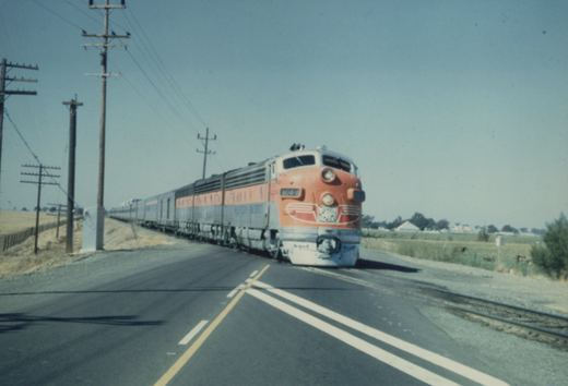 A Western Pacific orange and silver diesel locomotive numbered 904-B crosses a paved road with a line of matching passenger cars behind it, under a clear blue sky.