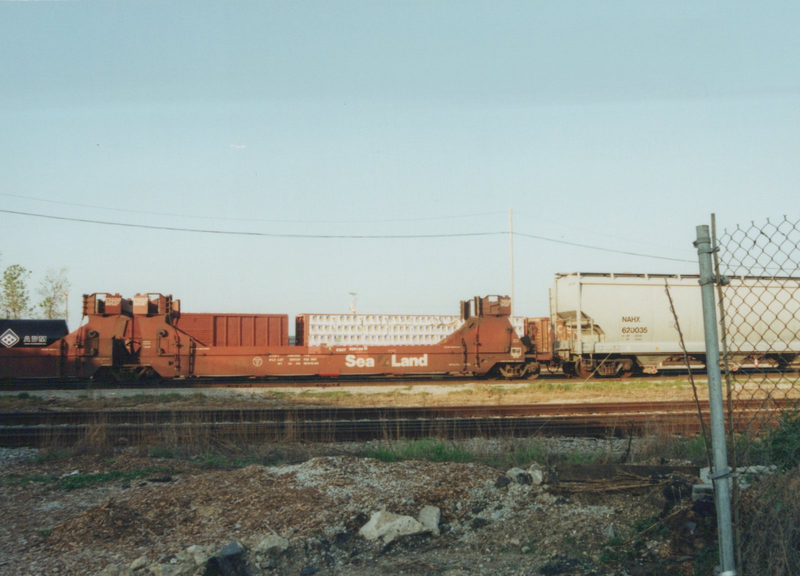 Side view of a freight train with a CSX flatcar carrying containers, an ADM tank car, and a NAHX covered hopper, on a sunny day beside a chain-link fence.