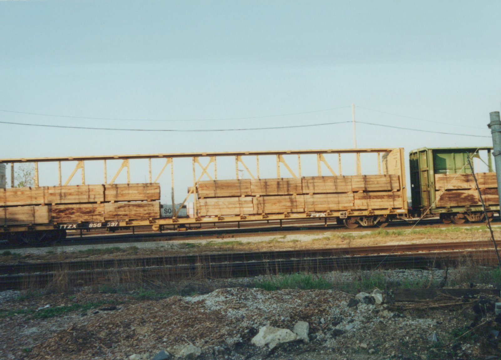 A side view of a freight train carrying lumber on a railroad main line, with visible TTX reporting marks and flatcars.