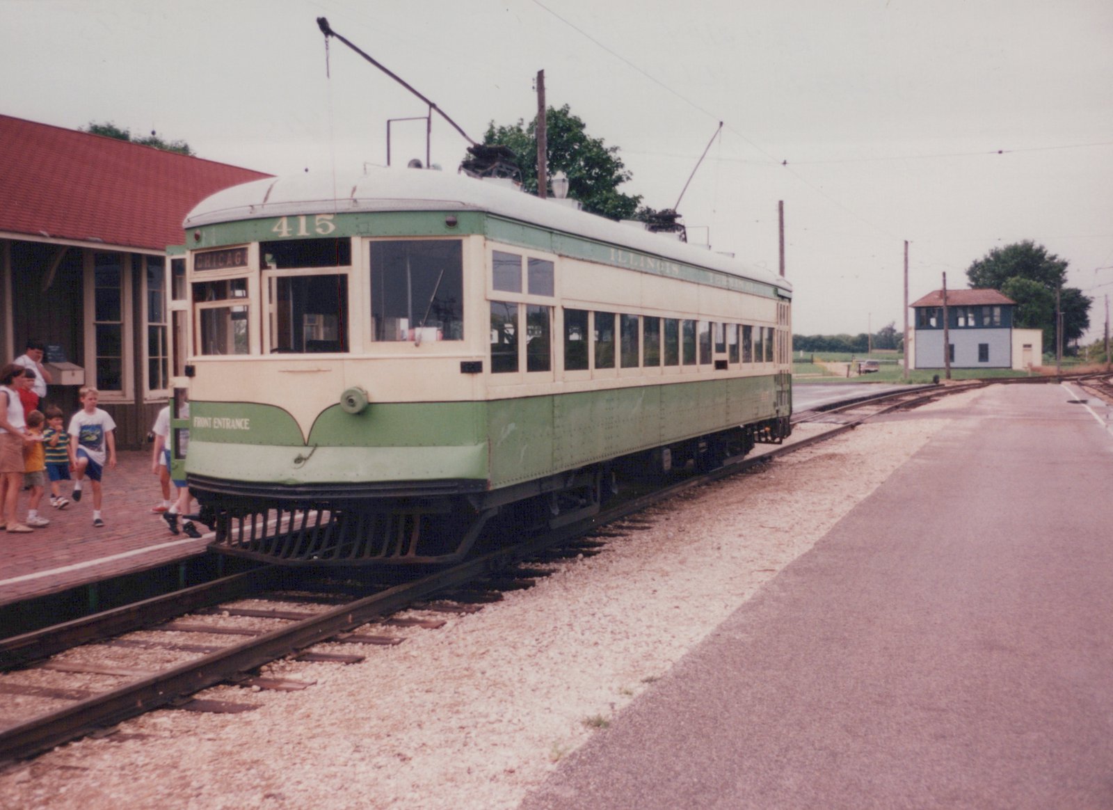 A green and cream interurban car numbered 415, lettered for the Illinois Terminal Railroad, is parked at a station platform with people nearby and overhead wires above.
