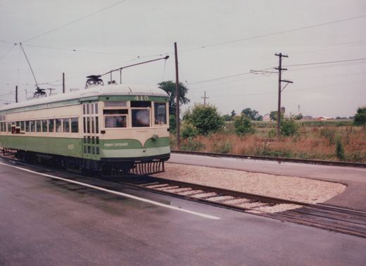 A vintage green and cream interurban car numbered 415 is on a track next to a paved road, with overhead wires and open farmland in the background.