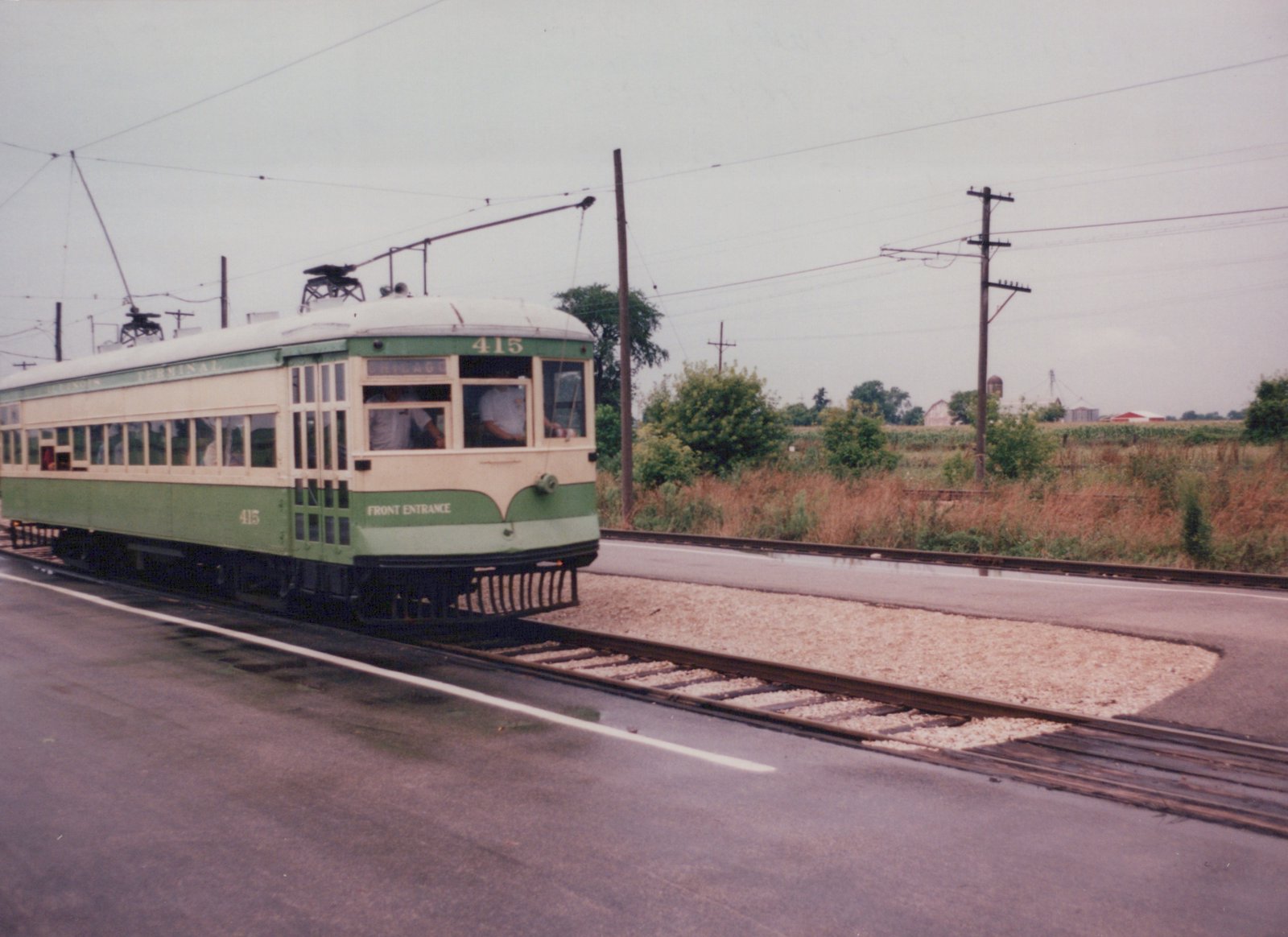 A vintage green and cream interurban car numbered 415 is on a track next to a paved road, with overhead wires and open farmland in the background.