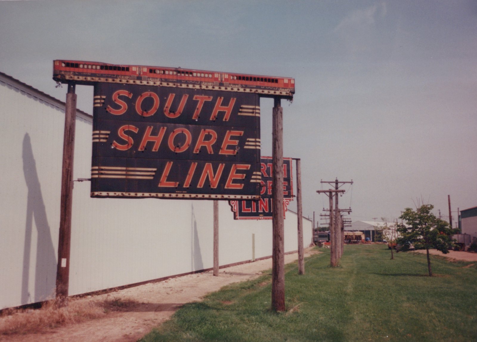 A large sign reading 'SOUTH SHORE LINE' with a red train graphic on top, mounted on wooden poles next to a white building and a grassy area where utility poles line the path.