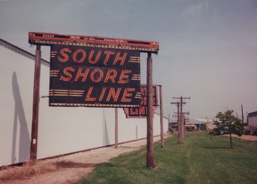 A large sign reading 'SOUTH SHORE LINE' with a red train graphic on top, mounted on wooden poles next to a white building and a grassy area where utility poles line the path.