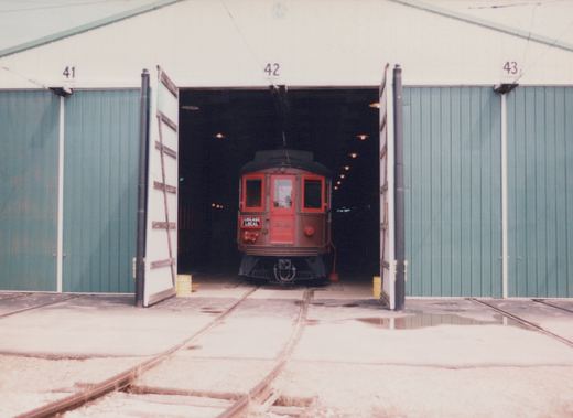 Front view of a red and black Birney interurban car marked 'CHICAGO LOCAL' inside a carbarn, with the car partially in shadow and tracks visible on the ground.