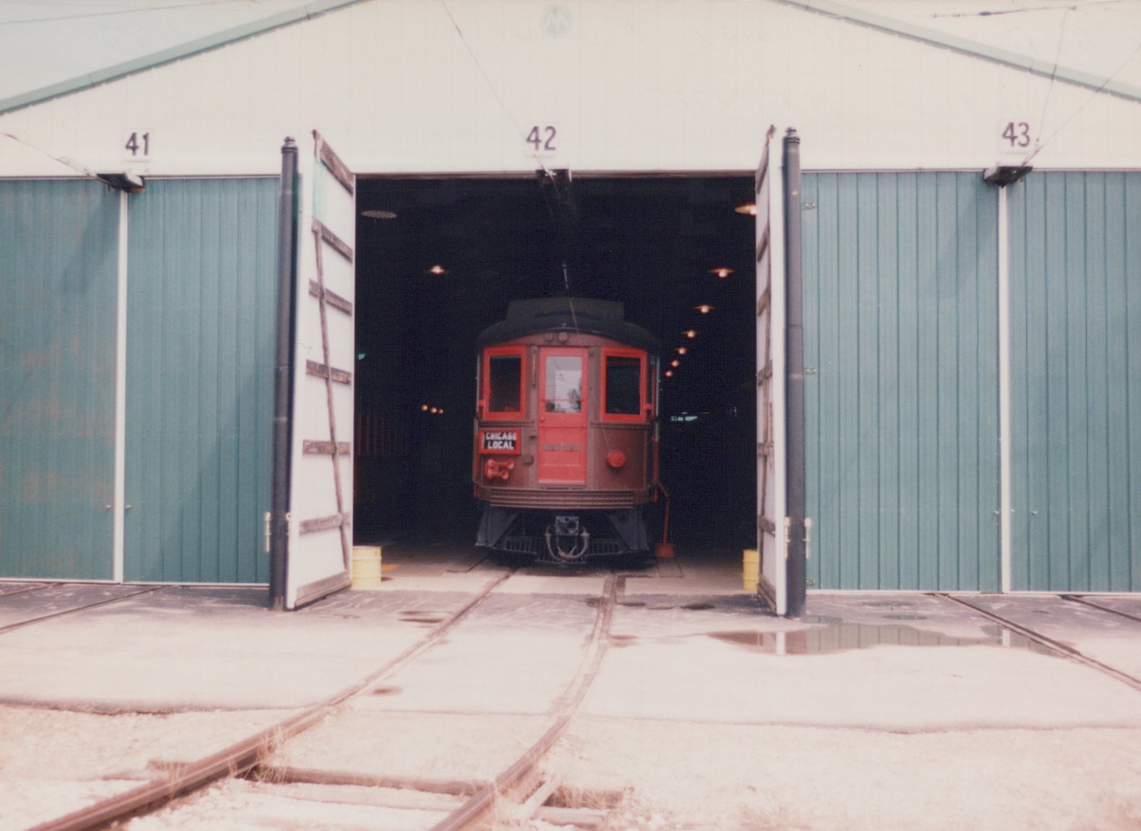 Front view of a red and black Birney interurban car marked 'CHICAGO LOCAL' inside a carbarn, with the car partially in shadow and tracks visible on the ground.