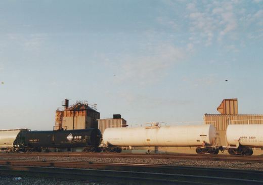 Side view of three freight tank cars on railroad tracks with industrial buildings in the background under a blue sky.