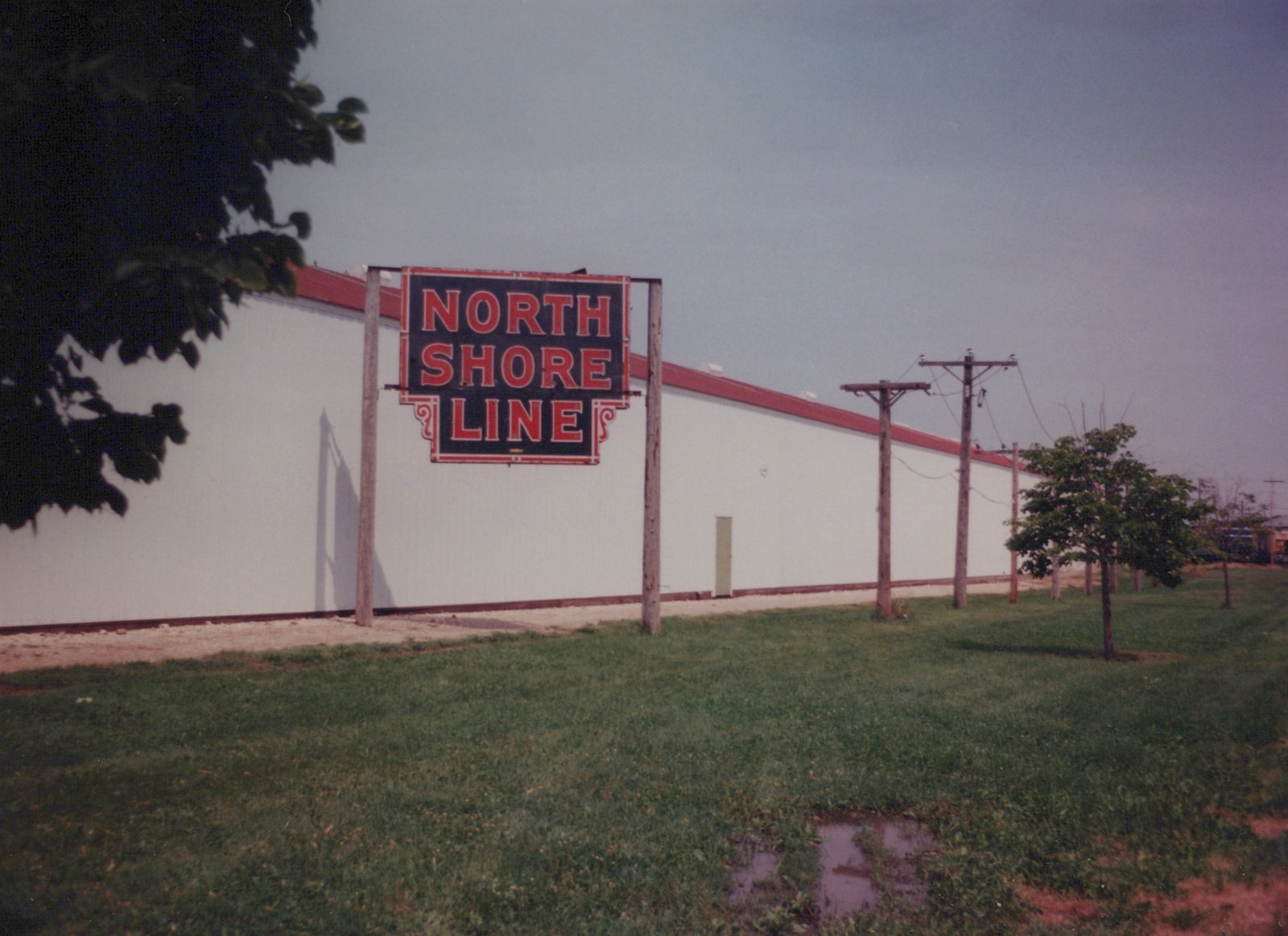 A large red and black sign reading 'NORTH SHORE LINE' is mounted on wooden posts in front of a long, white industrial building with a red roof trim, surrounded by grass and utility poles under an overcast sky.