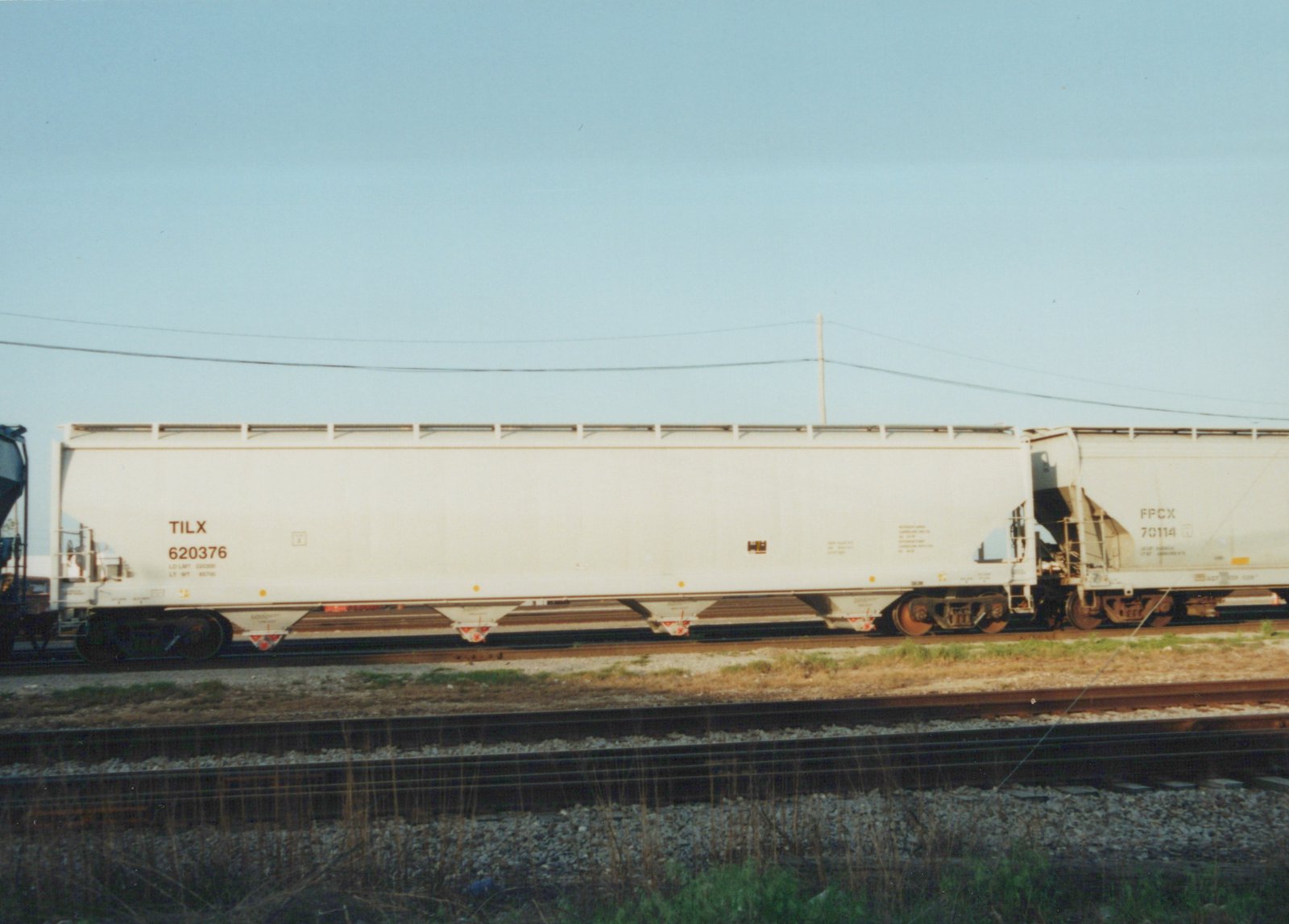 Side view of a white TILX covered hopper car, number 620376, coupled to an FPCX car, number 70114, sitting on railroad tracks with a clear blue sky above.