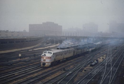 A white and orange Chicago & North Western EMD F7 locomotive, numbered 127, pulls a train through a multi-track rail yard with industrial buildings in the foggy background.
