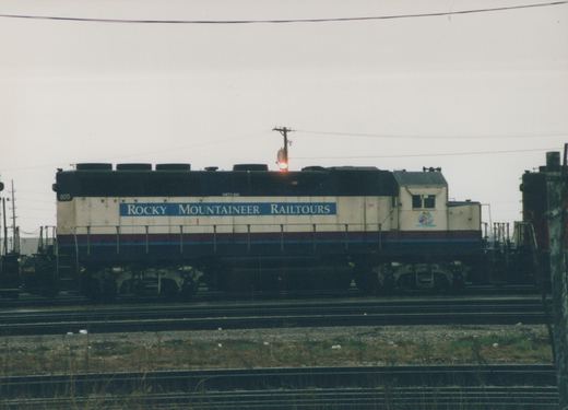 Side view of a cream-colored diesel locomotive with a blue and red stripe, lettered 'Rocky Mountaineer Railtours' and numbered 805, parked on multiple tracks in a rail yard under an overcast sky.