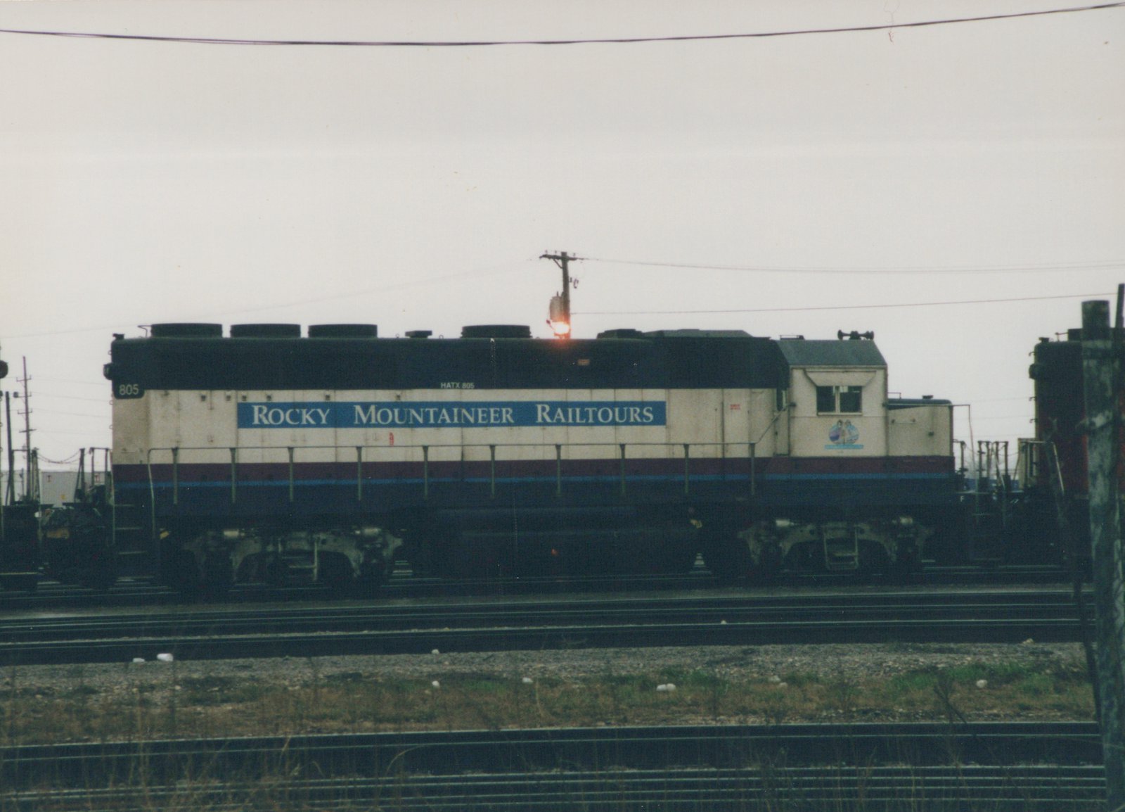 Side view of a cream-colored diesel locomotive with a blue and red stripe, lettered 'Rocky Mountaineer Railtours' and numbered 805, parked on multiple tracks in a rail yard under an overcast sky.