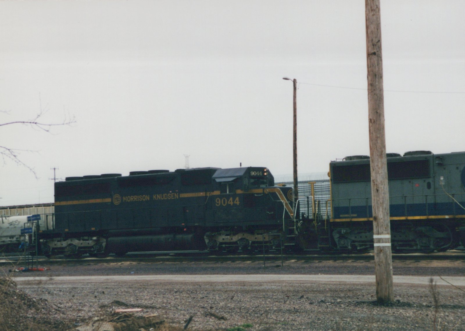 A dark green Morrison Knudsen diesel locomotive with the number 9044 is sitting on tracks in a gravel yard under an overcast sky, with another locomotive partially visible behind it.