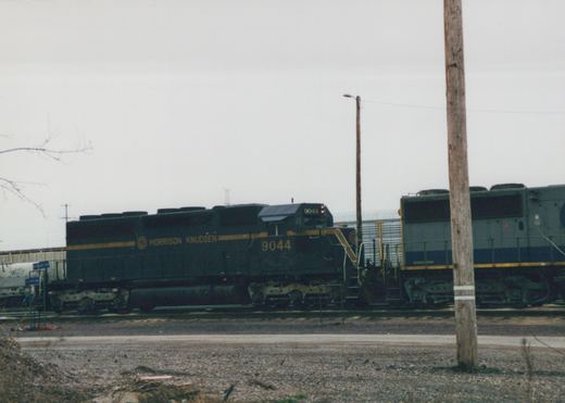A dark green Morrison Knudsen diesel locomotive with the number 9044 is sitting on tracks in a gravel yard under an overcast sky, with another locomotive partially visible behind it.