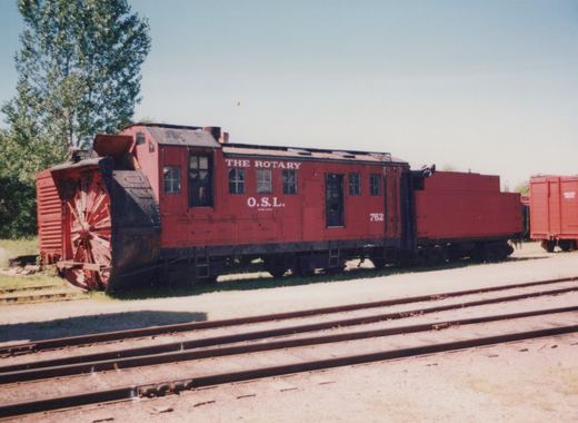 A red rotary snowplow with 'O.S.L.' and number '762' lettered on its side sits on railroad tracks in a sunny yard with trees in the background.