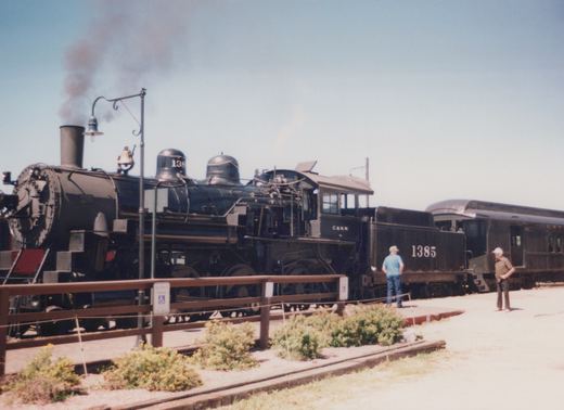 A black steam locomotive numbered 1385 with C&NW lettering is parked on tracks behind a wooden fence, with two men standing nearby on a sunny day.