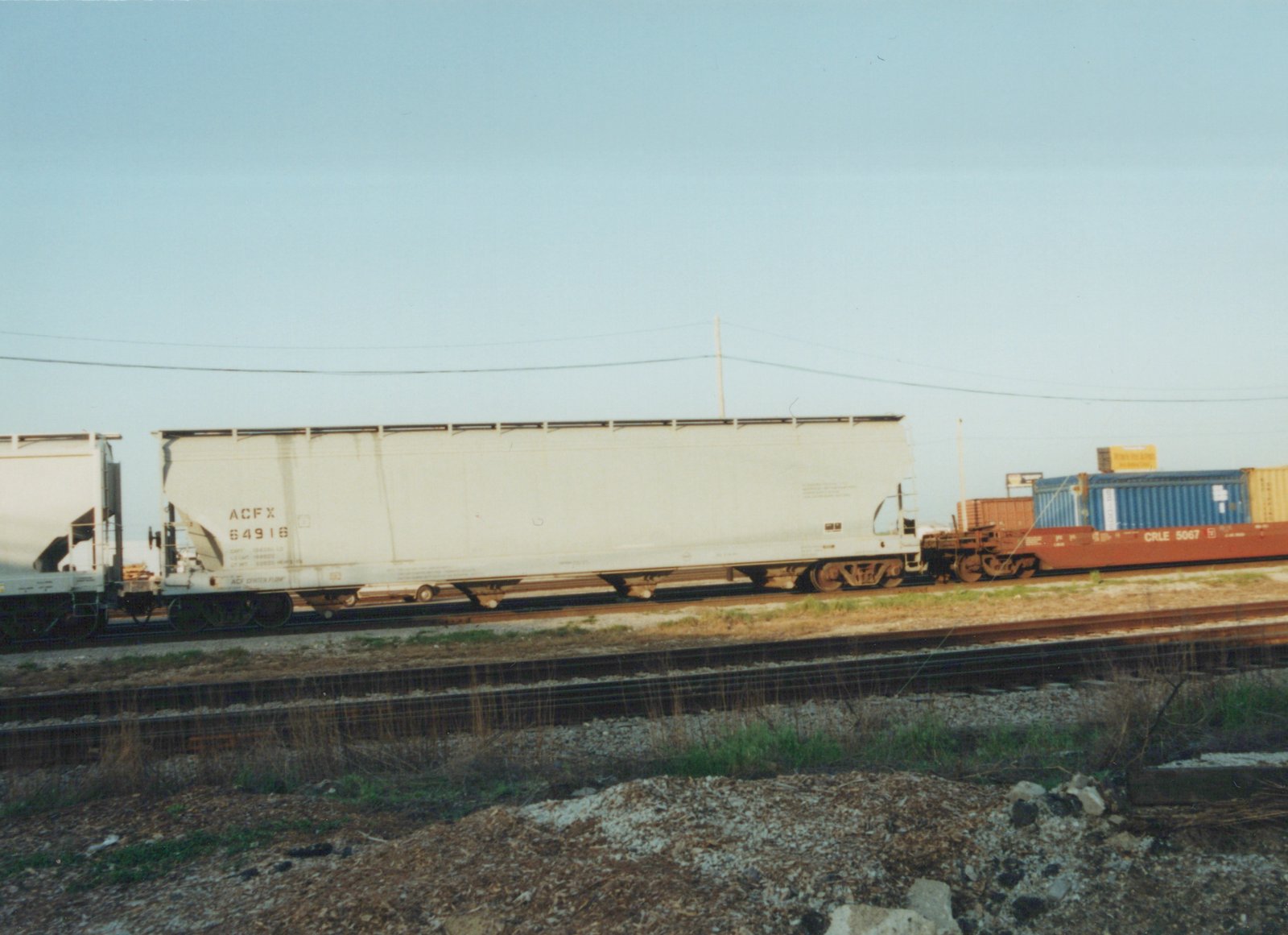 Side view of a freight train on a railroad main line under a clear sky, with a white ACF Industries gondola car and a red flatcar carrying blue and yellow shipping containers.