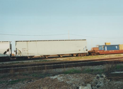 Side view of a freight train on a railroad main line under a clear sky, with a white ACF Industries gondola car and a red flatcar carrying blue and yellow shipping containers.