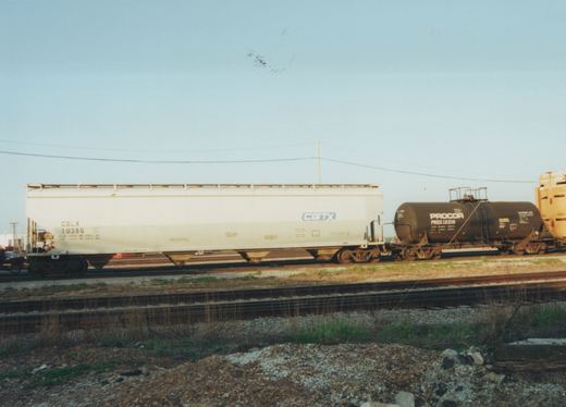 A white freight car with a CGTX logo and a black tank car with PROCOR lettering are parked side by side on a railroad main line, with tracks, gravel and dry grass in the foreground.