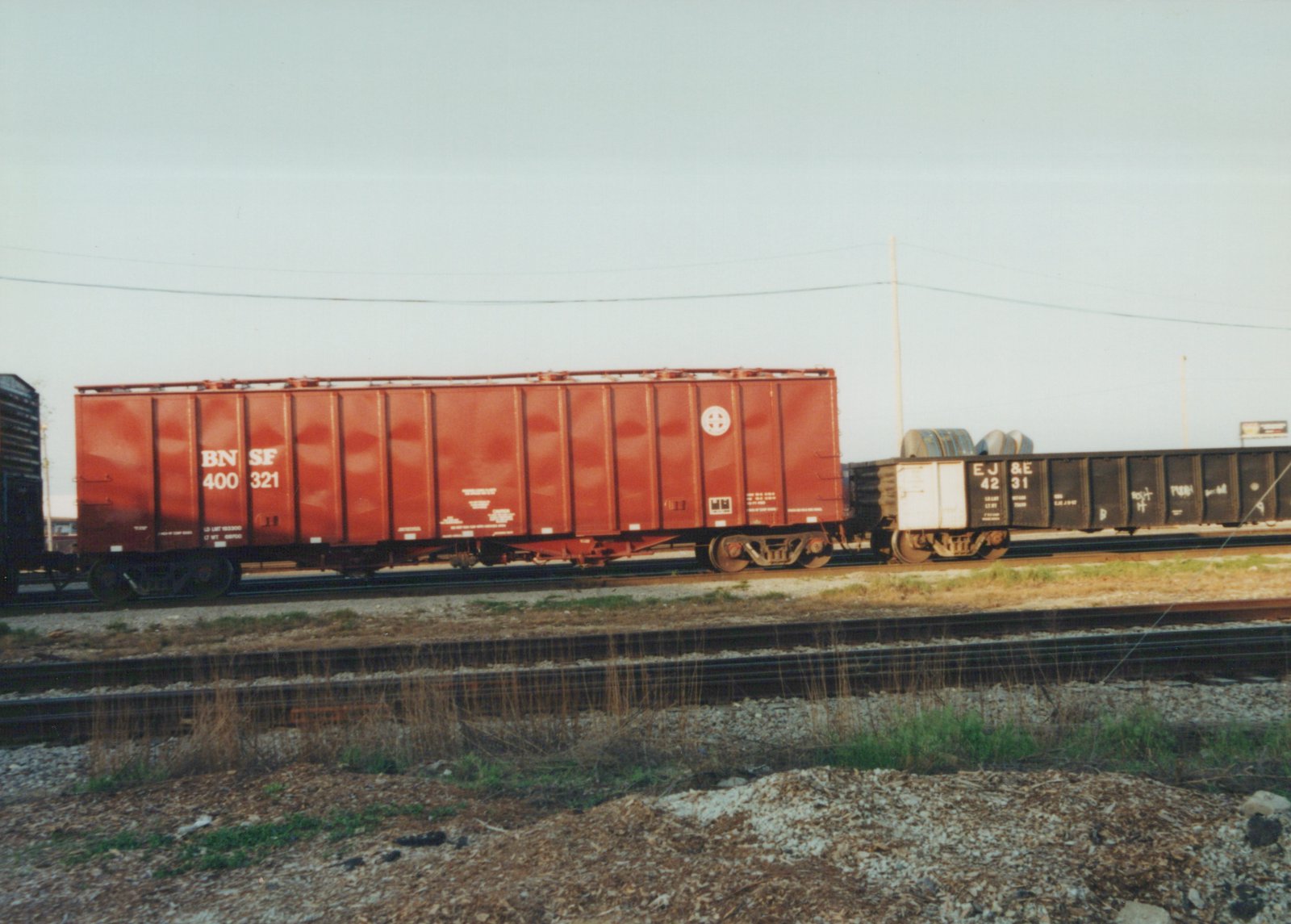 Side view of two freight cars on a railroad track: a red Burlington Northern hopper followed by a black Elgin Joliet and Eastern gondola, with a clear sky and grassy ground in the foreground.