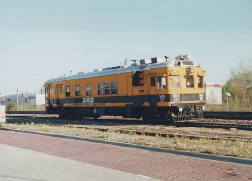 A yellow and black self-propelled railcar labeled 'SRS 137' and 'Sperry Rail Service' sits on tracks beside a paved walkway under a clear sky.