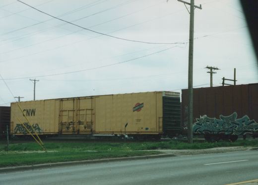 A yellow Chicago and North Western freight car and a brown Missouri Pacific car with blue graffiti sit on railroad tracks beside a road with grass and utility poles under a cloudy sky.