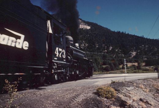 A black steam locomotive with the number 473 on its tender moves along a paved road next to a steep, tree-covered hillside on a sunny day.