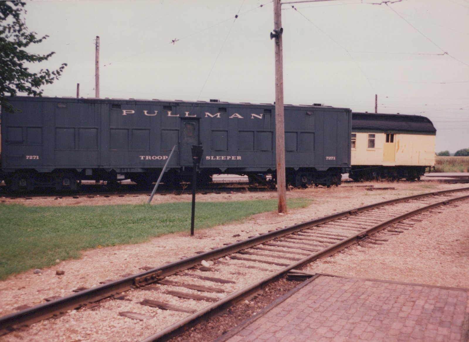 A dark green Pullman troop sleeper car numbered 7271 sits on tracks next to a yellow passenger car under overhead wires, with grass and a brick path in the foreground.