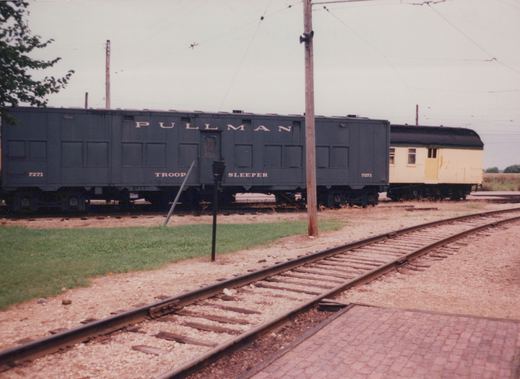 A dark green Pullman troop sleeper car numbered 7271 sits on tracks next to a yellow passenger car under overhead wires, with grass and a brick path in the foreground.