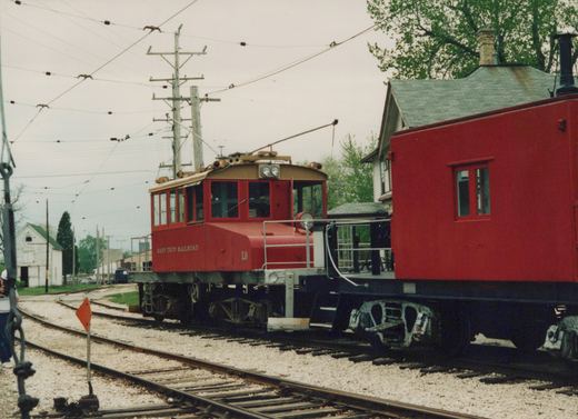 A red electric locomotive labeled East Troy Railroad L9 is shown on tracks beside a red caboose, with overhead wires and a house in the background.