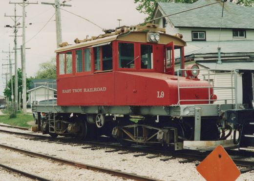 A red electric line car, number L9, for the East Troy Railroad, parked on tracks with overhead wires and a house in the background.