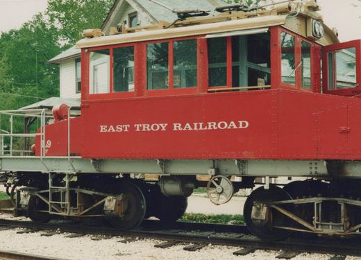 A bright red electric railcar with a cream-colored roof, marked 'EAST TROY RAILROAD' and numbered 'L9', is parked on railroad tracks beside a house.