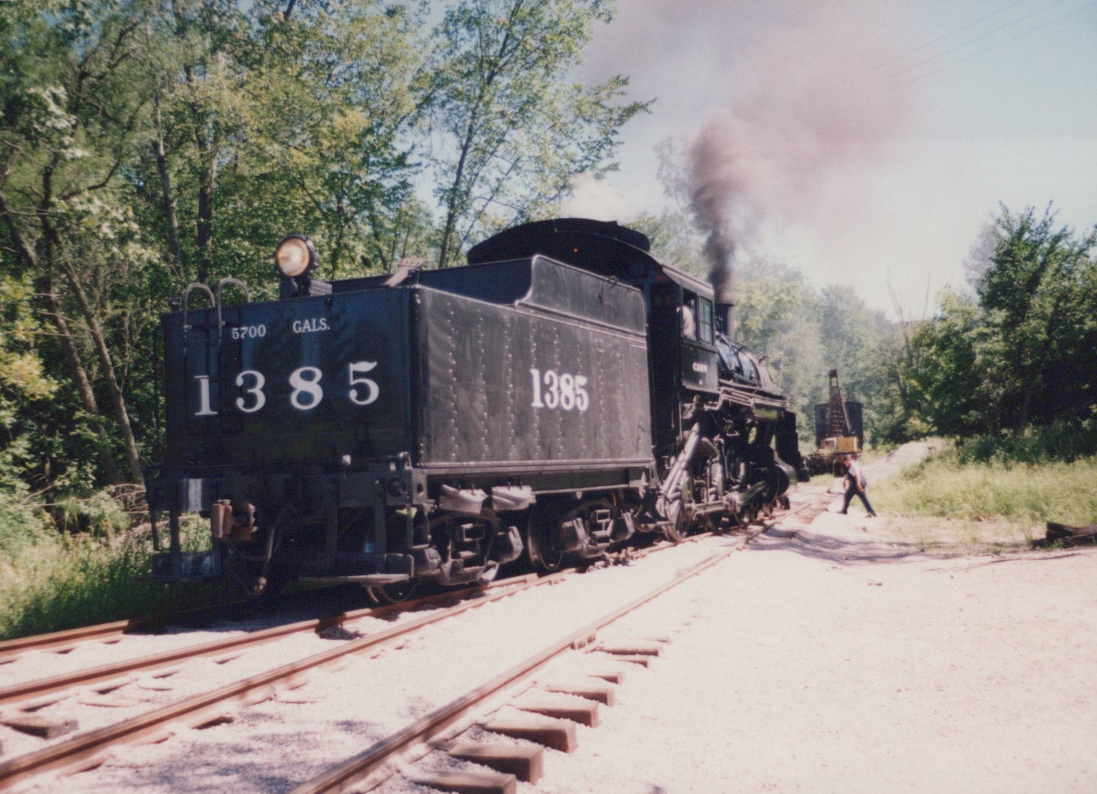 A black steam locomotive number 1385 with tender runs on a set of tracks surrounded by green trees on a sunny day, with one person walking nearby.