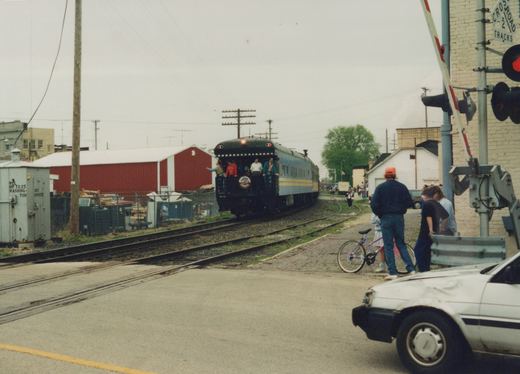 A dark caboose with a yellow stripe, marked CN and CM 98, moves along tracks past a railroad crossing where people stand watching; a white car is parked in the foreground and a red barn is in the background.