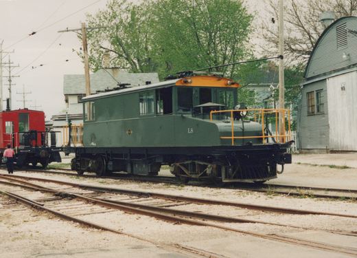 A green and orange electric locomotive numbered L8 stands on tracks in a yard with trees and a building nearby.