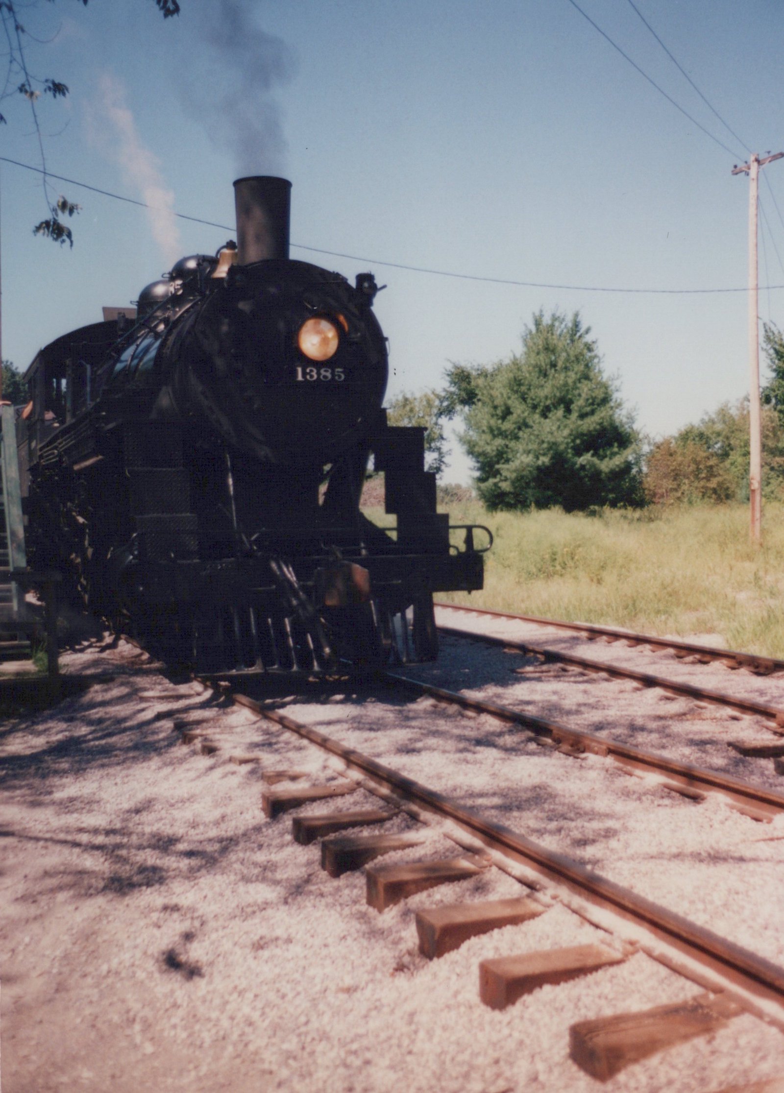 A black steam locomotive with the number 1385 on its front, on railroad tracks in a sunny, grassy area.