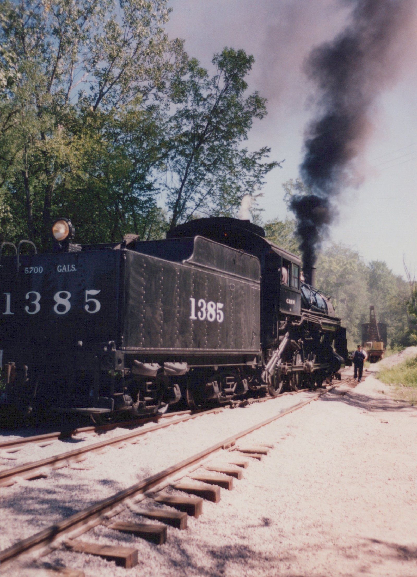 A black steam locomotive numbered 1385 with 'C&NW' lettering runs on a track, emitting black smoke, with green trees and a rail crane in the background.