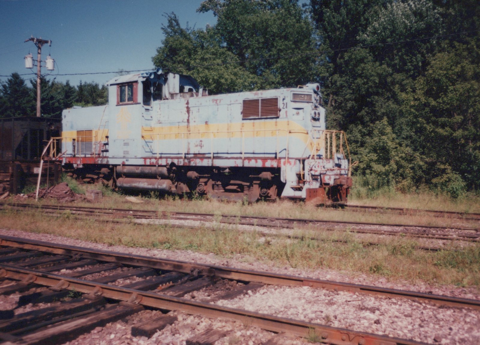 A side view of a weathered Wisconsin Central Ltd. diesel locomotive, number 21, painted in light blue with yellow and red accents, sitting on a set of tracks in a grassy yard with trees in the background.