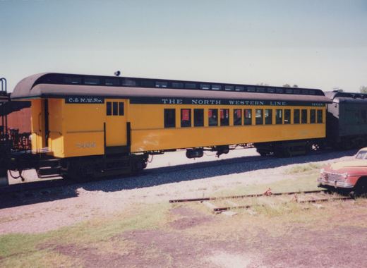 A bright yellow Chicago and North Western passenger car numbered 7409 sits on a track in a sunny yard, with a classic red and yellow car parked nearby and another dark railcar coupled behind it.