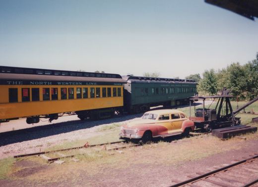 Two vintage Chicago and North Western passenger cars, one yellow and one green, sit on parallel tracks in a grassy yard on a sunny day; a 1940s car is mounted on a rail trailer nearby.