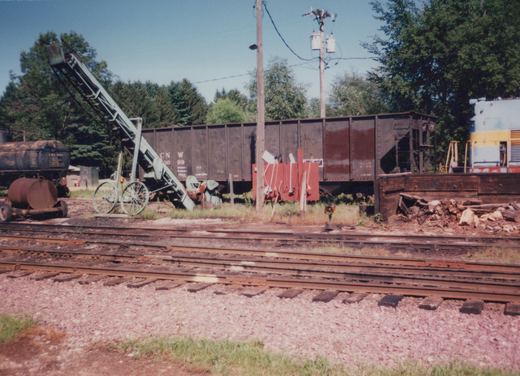 A sunlit railroad yard scene with two freight cars on parallel tracks, a wooden loading conveyor angled towards one car, a small tank trailer, and a pile of wood debris, set against a backdrop of dense trees.