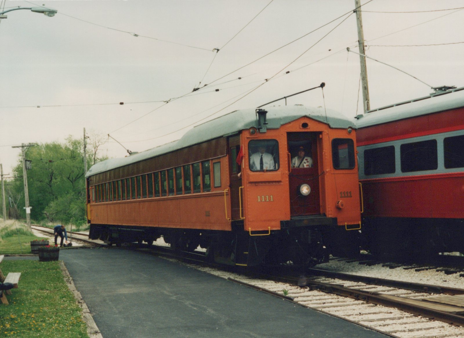An orange and green interurban car numbered 1111 is stopped on tracks beside a paved path, with two crew members visible in the cab and another person working on the track nearby.
