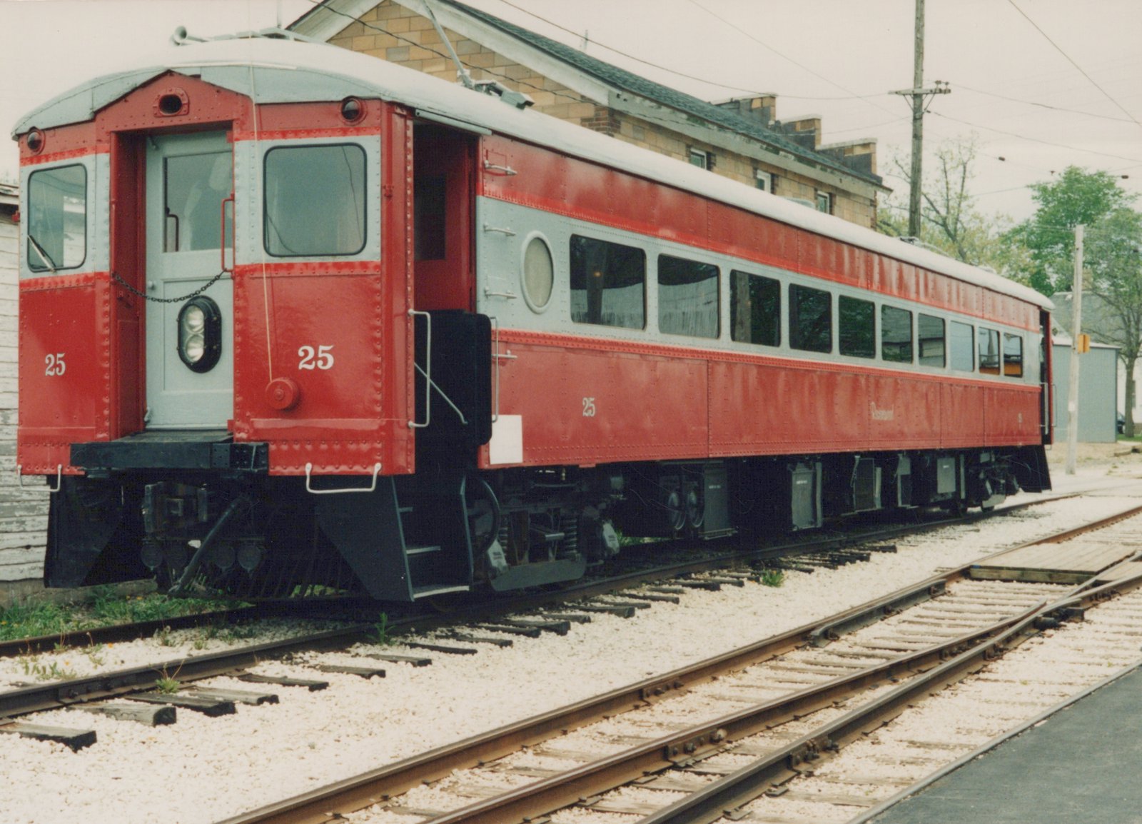 A red and silver interurban railcar with the number 25 and the name 'Rosemont' on its side, parked on train tracks in front of a brick building under an overcast sky.