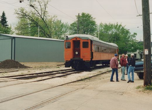 An orange and gray interurban car numbered 1111 sits on tracks next to a street, with four people standing nearby and a utility pole with a high voltage sign.