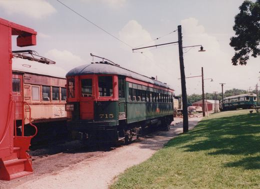 A vintage red and green interurban car numbered 715 is parked on a rail yard beside another historic car, with a modern green and white train visible in the background.
