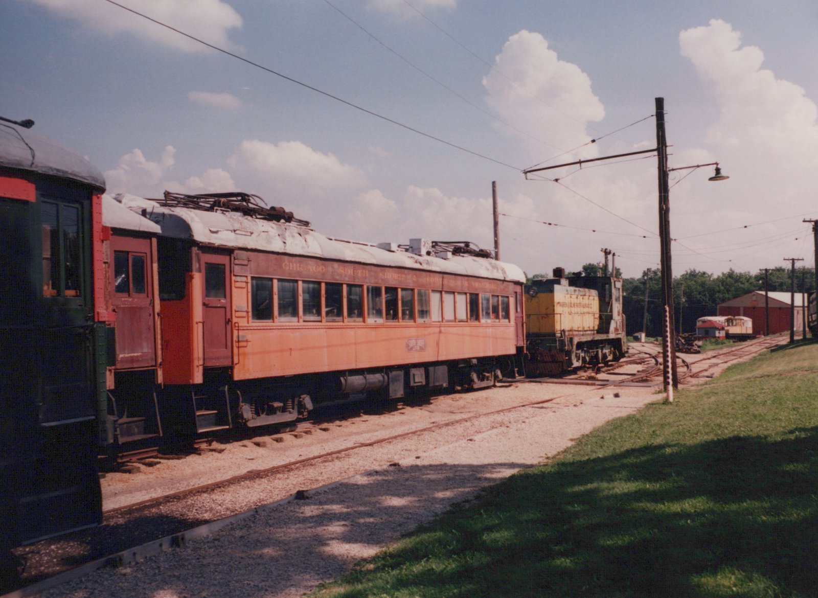 An orange interurban railcar and a yellow electric locomotive are parked side by side on parallel tracks in a grassy yard under a blue sky with white clouds.