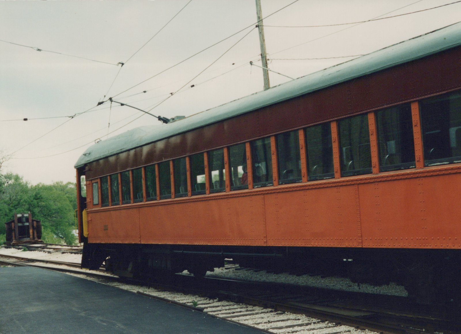 A side view of a vintage red-orange interurban car with a green roof, sitting on tracks under overhead wires on an overcast day.