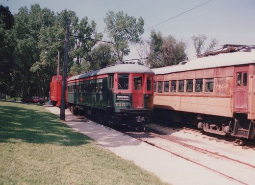 A green and red historic interurban car numbered 715 sits on a track next to a rusty red interurban car, with a red boxcar and a sedan in the background on a sunny day.