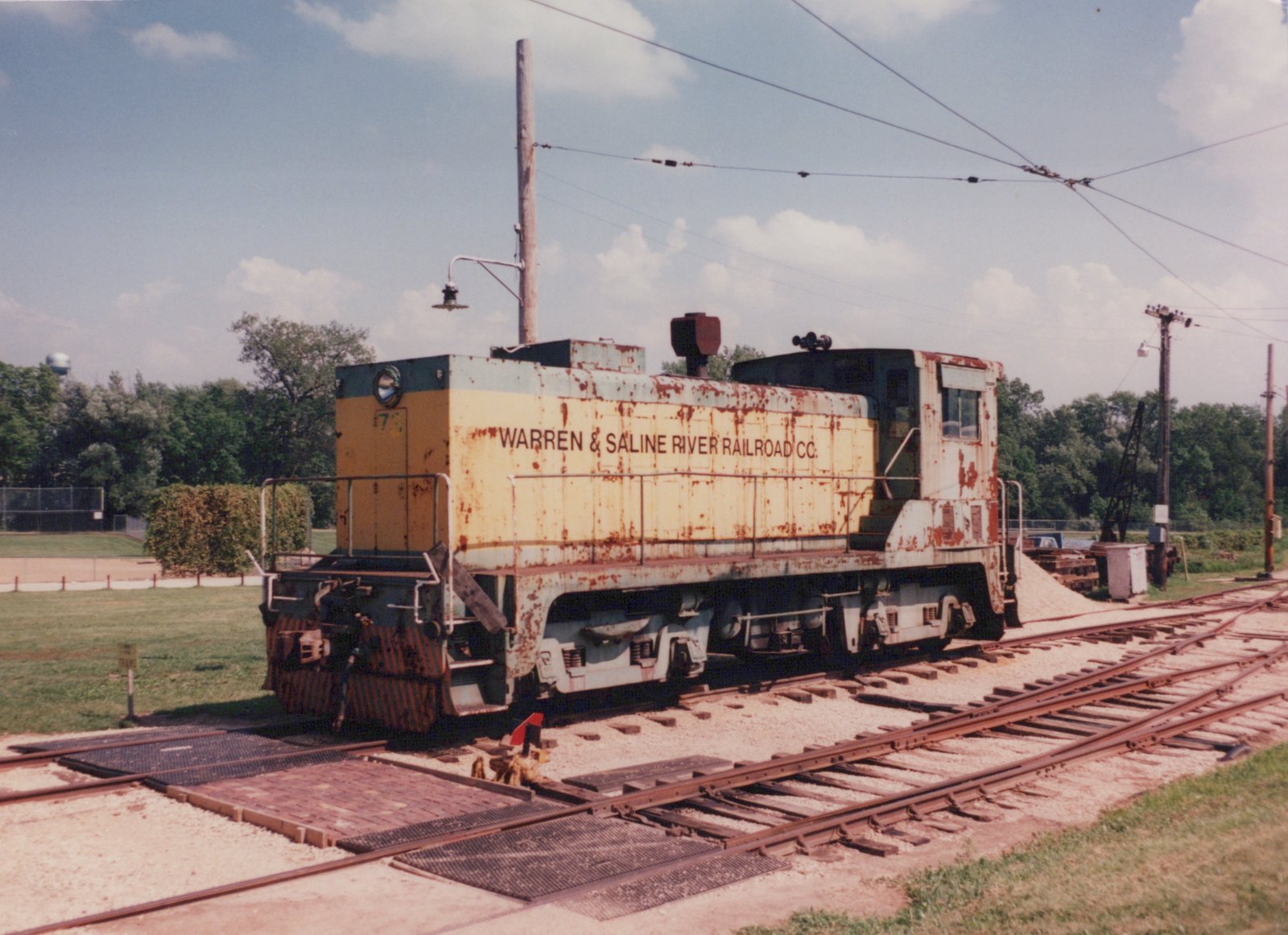 A rusty, yellow and green electric locomotive with 'WARREN & SALINE RIVER RAILROAD CO.' lettered on its side, marked with the number 7?, is parked on multiple tracks under overhead wires on a clear summer day.